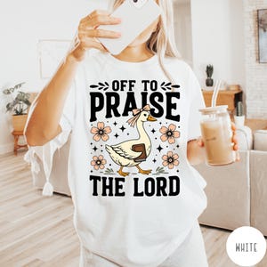 Puede incluir: Camiseta blanca con el texto "OFF TO PRAISE THE LORD". El gr&aacute;fico presenta un ganso de dibujos animados con un lazo, sosteniendo un libro, rodeado de flores y estrellas. La camiseta es de estilo c&oacute;modo e informal.