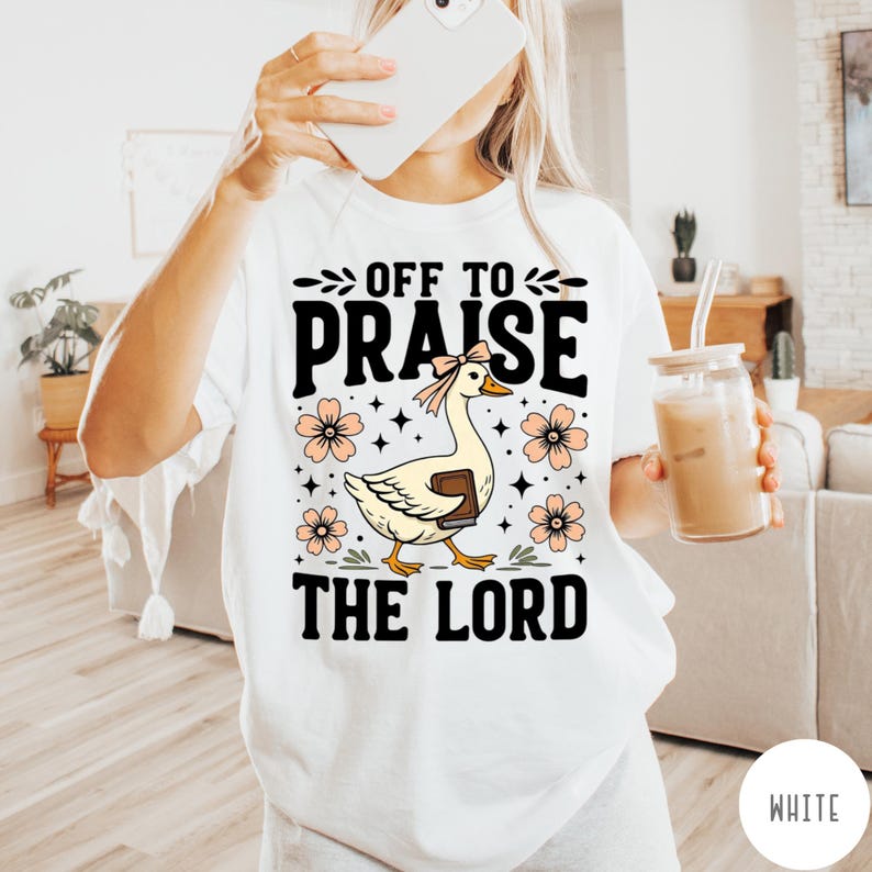 Puede incluir: Camiseta blanca con el texto "OFF TO PRAISE THE LORD". El gr&aacute;fico presenta un ganso de dibujos animados con un lazo, sosteniendo un libro, rodeado de flores y estrellas. La camiseta es de estilo c&oacute;modo e informal.