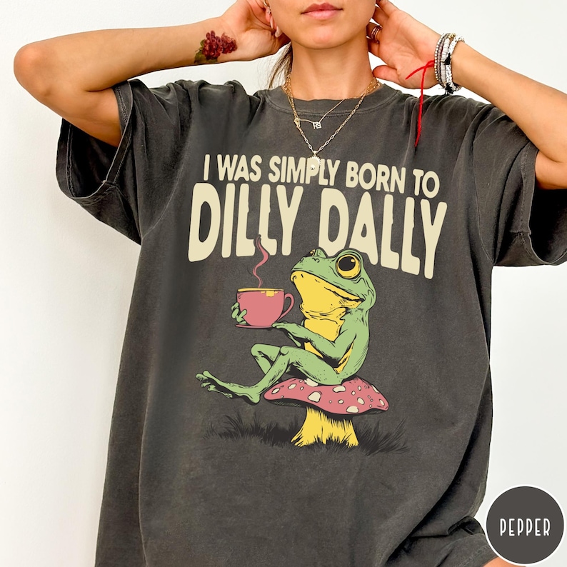 May include: Dark gray t-shirt with the phrase "I WAS SIMPLY BORN TO DILLY DALLY" in cream-colored text. A cartoon frog is seated on a mushroom, holding a pink teacup. The frog is green and yellow.
