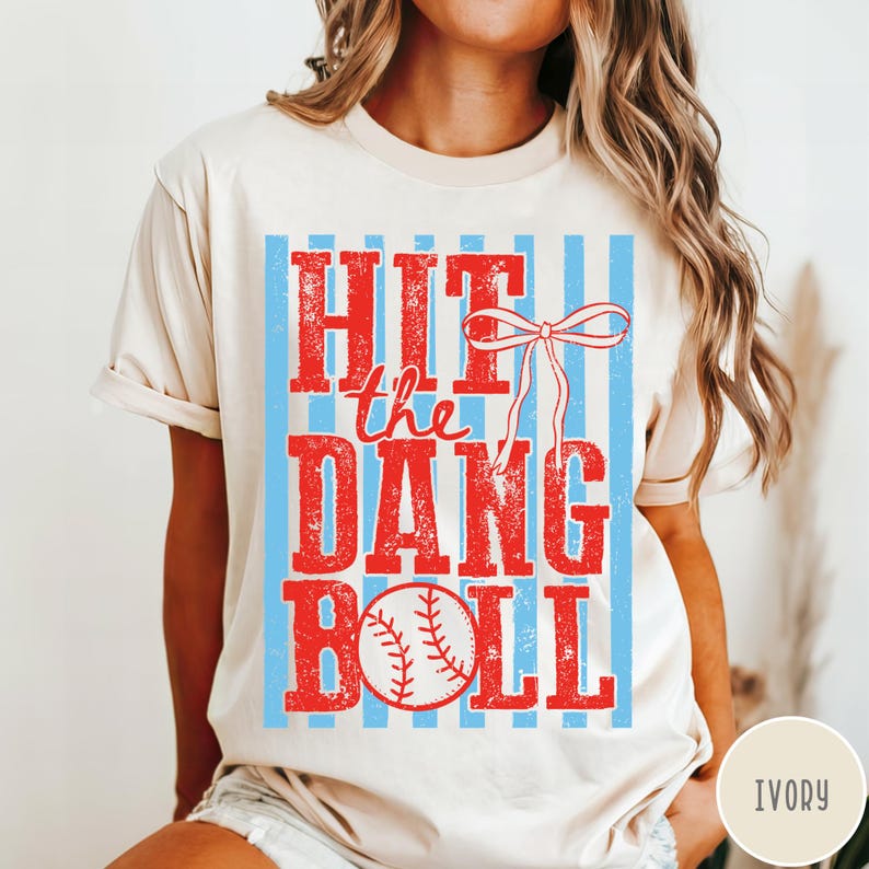 Pu&ograve; includere: T-shirt color avorio con grafica rossa e blu invecchiata. Il design presenta la scritta "HIT the DANG BALL" sopra un'illustrazione di baseball. Strisce verticali blu formano uno sfondo per il testo e la palla da baseball.