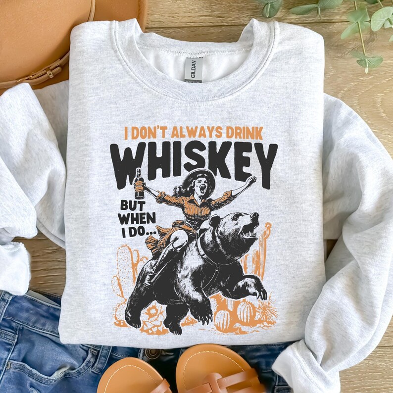 May include: A gray sweatshirt with a graphic print of a woman riding a bear. The text on the shirt reads "I don't always drink whiskey, but when I do..."