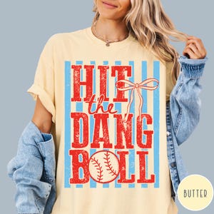 Pu&ograve; includere: T-shirt color crema con la scritta "HIT the DANG BALL" in rosso, con una grafica di baseball. Il design include strisce verticali blu e un fiocco. La parola "BUTTER" &egrave; in un cerchio in basso a destra.