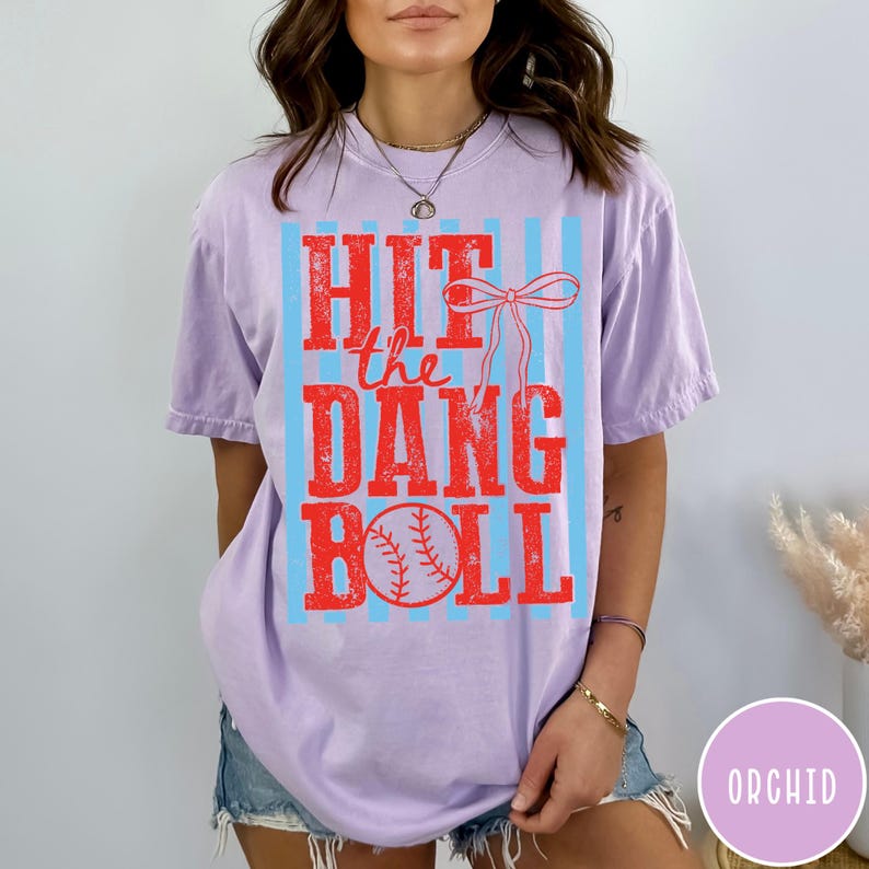 Pu&ograve; includere: Una maglietta lilla con la scritta rossa "HIT the DANG BALL" su sfondo a righe azzurre. Un grafico di baseball rosso in basso. La maglietta &egrave; in stile casual.