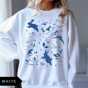 May include: White sweatshirt with a repeating pattern of blue rabbits, planets, and stars. The word "WHITE" is printed in a black box at the bottom. The design has a whimsical, celestial theme.