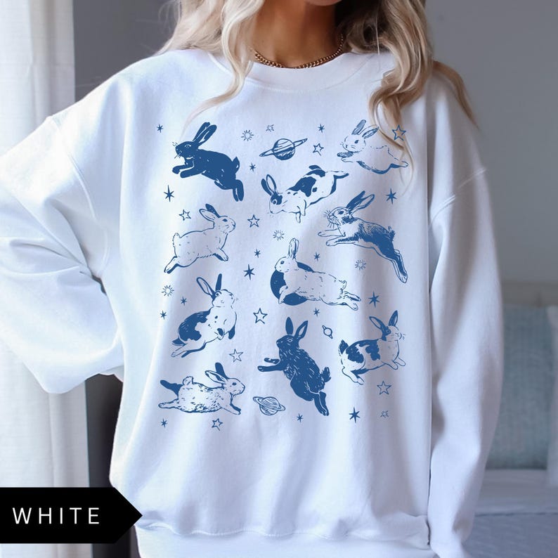 May include: White sweatshirt with a repeating pattern of blue rabbits, planets, and stars. The word "WHITE" is printed in a black box at the bottom. The design has a whimsical, celestial theme.