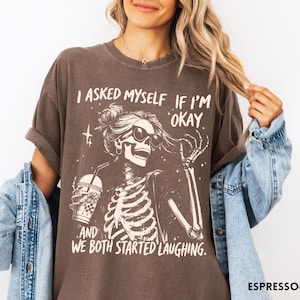 May include: Espresso-colored t-shirt featuring a skeleton graphic wearing sunglasses and holding a drink. The text reads, "I asked myself if I'm okay, and we both started laughing."