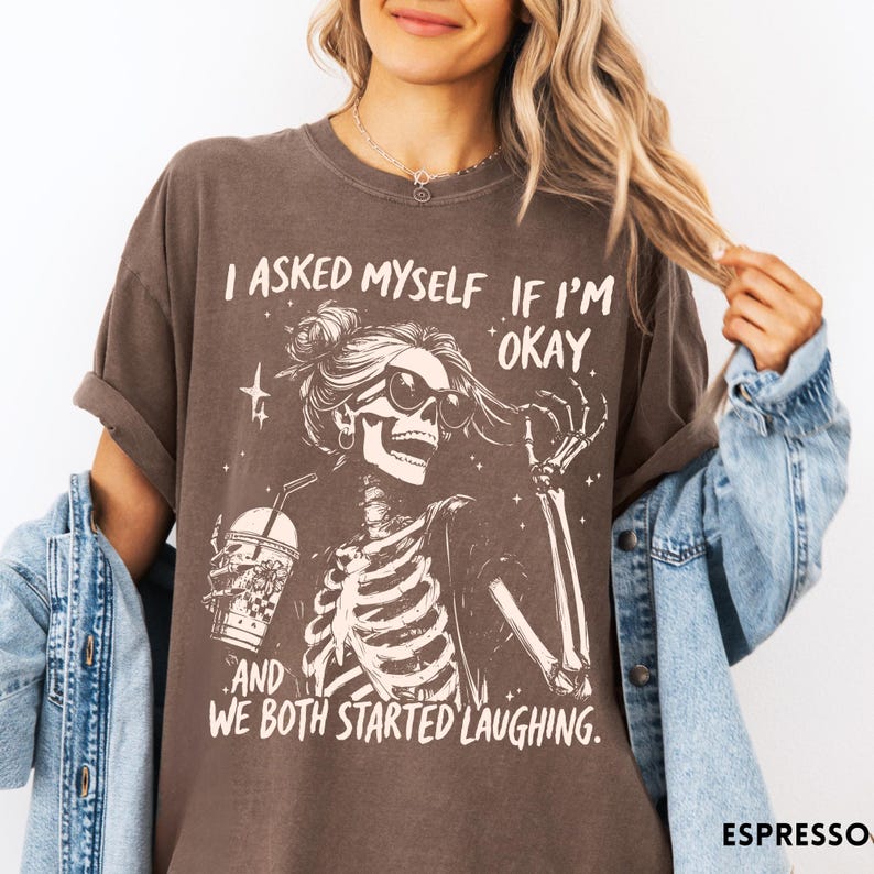 May include: Espresso-colored t-shirt featuring a skeleton graphic wearing sunglasses and holding a drink. The text reads, "I asked myself if I'm okay, and we both started laughing."