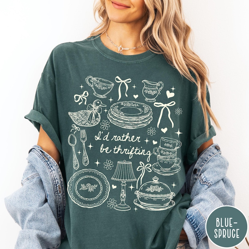 May include: Dark teal t-shirt featuring a white illustration of vintage crockery, including plates, teacups, and a duck ornament. The text "I'd rather be thrifting" is below the design. The shirt has rolled-up sleeves.