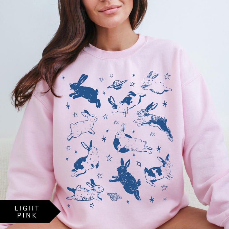 May include: Pale pink sweatshirt featuring a navy blue rabbit and space-themed design. The graphic includes rabbits, stars, and planets. The text "LIGHT PINK" is printed in a black box at the bottom.