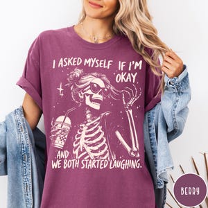 May include: Berry-colored t-shirt featuring a skeleton graphic with sunglasses and a drink. The text reads, "I asked myself if I'm okay, and we both started laughing." The shirt has short sleeves and a relaxed fit.