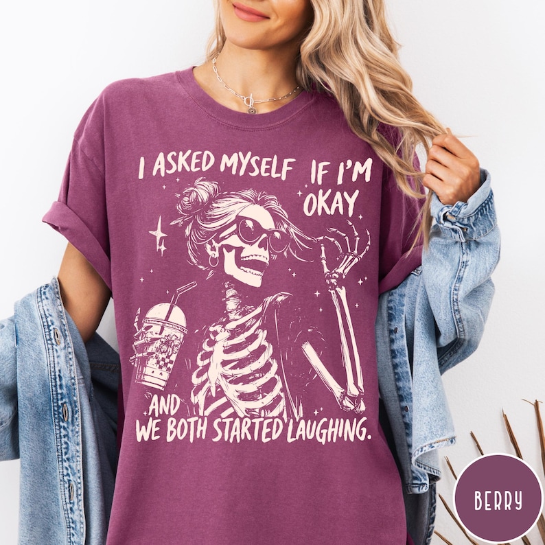 May include: Berry-colored t-shirt featuring a skeleton graphic with sunglasses and a drink. The text reads, "I asked myself if I'm okay, and we both started laughing." The shirt has short sleeves and a relaxed fit.