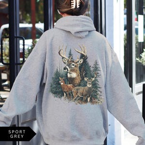 Op de afbeelding: Sportgrijze hoodie met een gedetailleerde afbeelding van herten in een bos. Het ontwerp omvat een hert met groot gewei, twee andere herten en groenblijvende bomen. De hoodie heeft een neutrale grijze kleur.