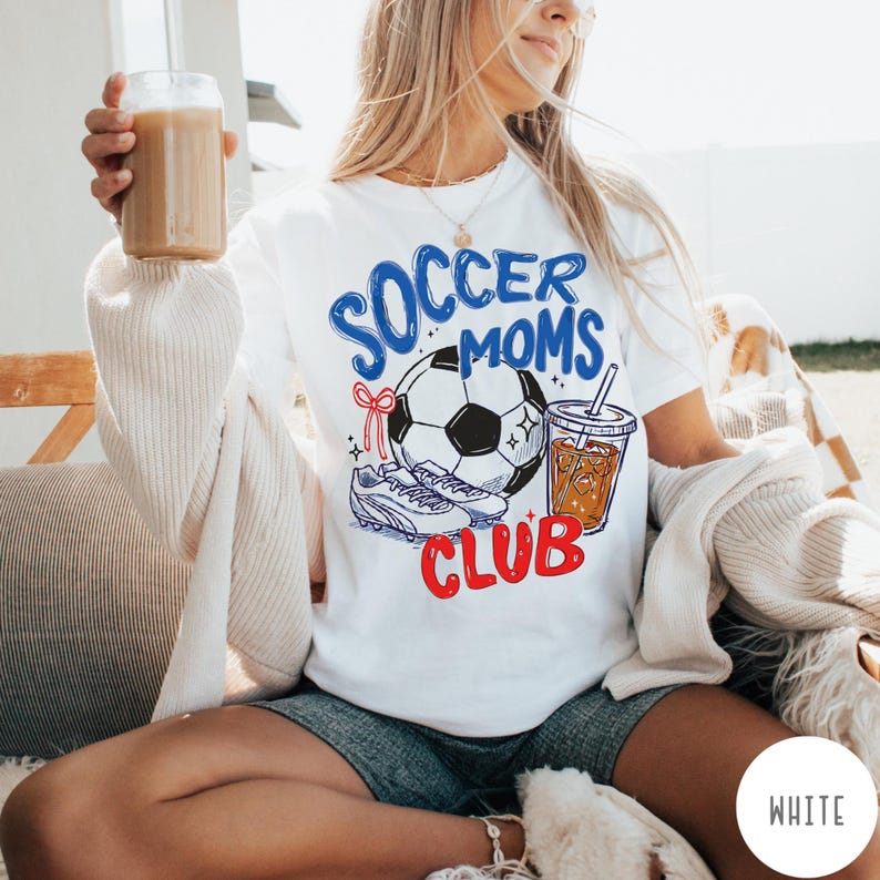 May include: White t-shirt with a graphic design featuring a soccer ball, cleats, and a beverage. The text on the shirt reads "Soccer Moms Club" in blue and red. The shirt is worn by a person.