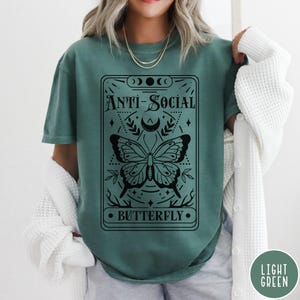 Puede incluir: Una camiseta verde azulada con un gr&aacute;fico negro de una mariposa y el texto "Anti-Social Butterfly".