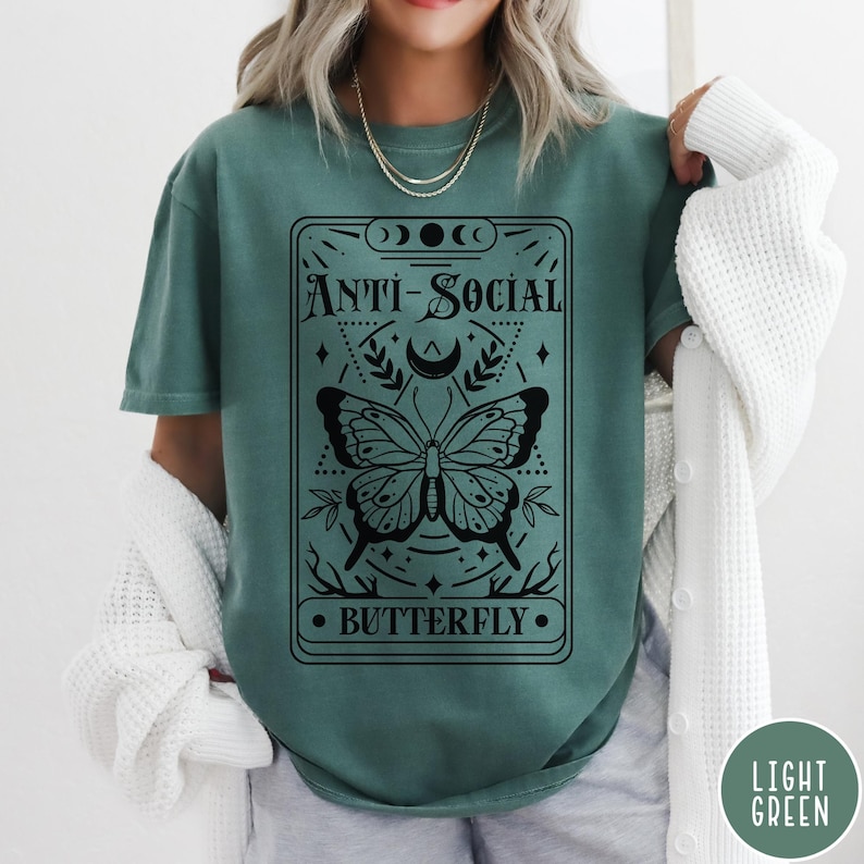 Puede incluir: Una camiseta verde azulada con un gr&aacute;fico negro de una mariposa y el texto "Anti-Social Butterfly".