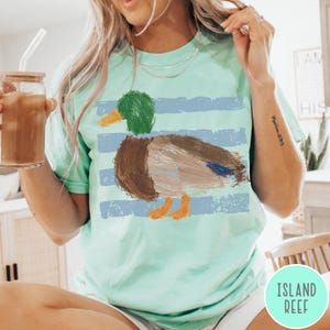 May include: A mint green short-sleeve t-shirt featuring a graphic of a mallard duck with a green head, brown body, and orange feet. The duck is layered over horizontal light blue distressed stripes. The shirt has a crew neckline and a loose fit.
