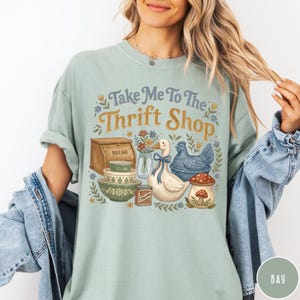 Op de afbeelding: Lichtgroen t-shirt met de tekst "Take Me To The Thrift Shop" in blauw en goud. De afbeelding bevat een brooddoos, kommen, een eend, een kip en een paddenstoel.