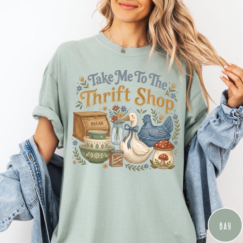 Op de afbeelding: Lichtgroen t-shirt met de tekst "Take Me To The Thrift Shop" in blauw en goud. De afbeelding bevat een brooddoos, kommen, een eend, een kip en een paddenstoel.