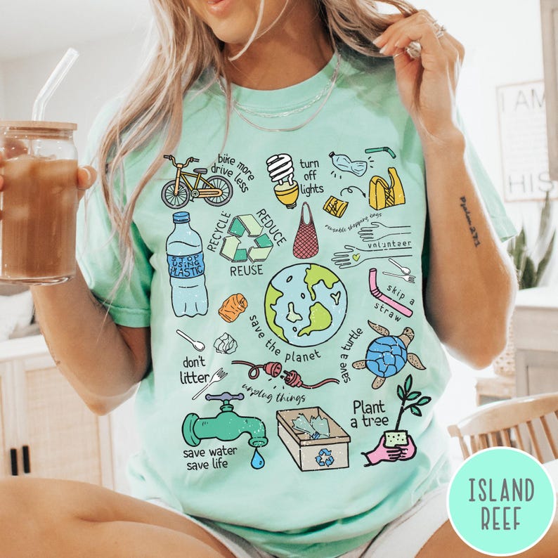 May include: A light blue t-shirt with a colourful graphic design featuring various eco-friendly tips and illustrations. The design includes a bicycle, a lightbulb, a reusable bag, a turtle, a tree, and the text "save the planet".