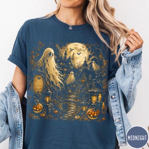 Puede incluir: Una camiseta azul oscuro con un gr&aacute;fico detallado en amarillo dorado de una escena embrujada con fantasmas, un castillo a la luz de la luna, un camino de piedra y calabazas talladas. El dise&ntilde;o incluye murci&eacute;lagos y una insignia MIDNIGHT.
