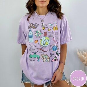 May include: A purple t-shirt featuring a graphic print of various illustrations related to environmentalism. The illustrations include a bicycle, a lightbulb, a water bottle, a recycling symbol, a globe, a turtle, a tree, and a faucet. The text on the shirt reads "bike more drive less", "turn off lights", "reduce", "recycle", "reuse", "don't litter", "save the planet", "save a turtle", "skip a straw", "plant a tree", and "save water save life".