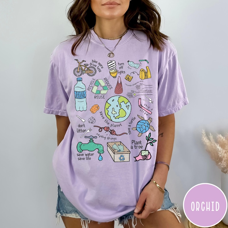 May include: A purple t-shirt featuring a graphic print of various illustrations related to environmentalism. The illustrations include a bicycle, a lightbulb, a water bottle, a recycling symbol, a globe, a turtle, a tree, and a faucet. The text on the shirt reads "bike more drive less", "turn off lights", "reduce", "recycle", "reuse", "don't litter", "save the planet", "save a turtle", "skip a straw", "plant a tree", and "save water save life".