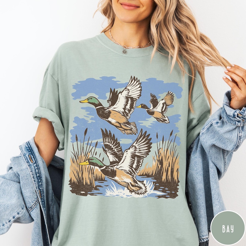 May include: Sage green t-shirt with a graphic of three ducks flying over water and reeds. The ducks are illustrated with brown, white, and green details against a blue sky. The shirt has a relaxed fit.