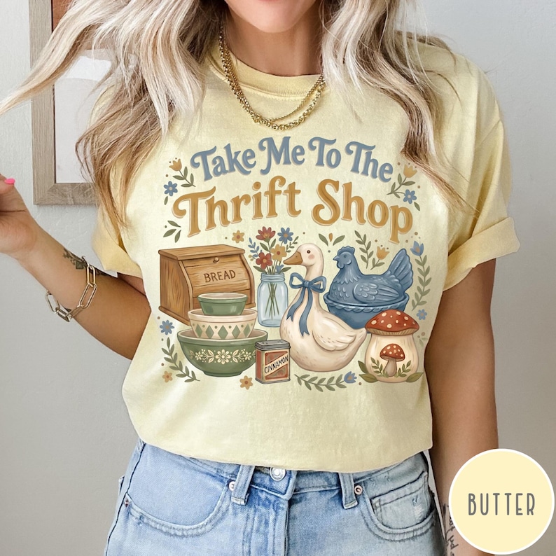 Op de afbeelding: Lichtgele t-shirt met de tekst "Take Me To The Thrift Shop" en een retro-ontwerp. Het ontwerp bevat een brooddoos, kommen, een eend, een kip en een paddenstoel, met bloemen en botanische details.