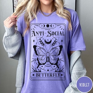 Puede incluir: Una camiseta morada con un dise&ntilde;o gr&aacute;fico negro que presenta una mariposa y el texto "Anti-Social Butterfly".
