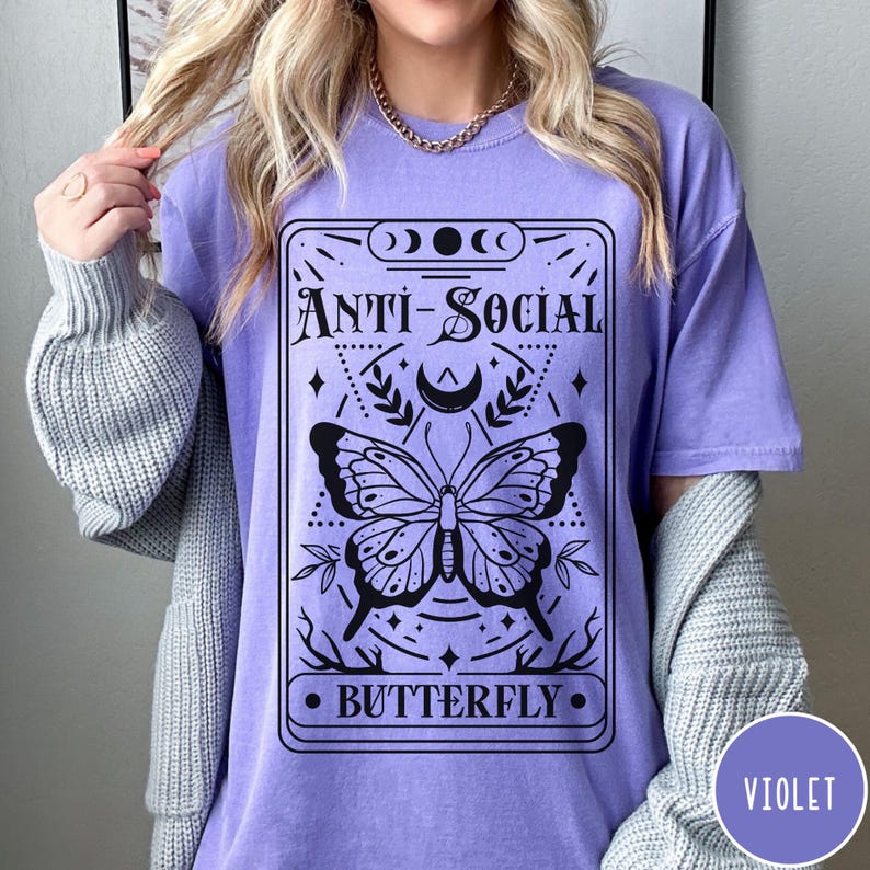 Puede incluir: Una camiseta morada con un dise&ntilde;o gr&aacute;fico negro que presenta una mariposa y el texto "Anti-Social Butterfly".