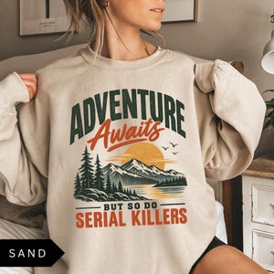 May include: A sand-colored crewneck sweatshirt featuring a graphic print with the text "ADVENTURE Awaits BUT SO DO SERIAL KILLERS". The design includes a mountain landscape, pine trees, a lake, and a sun in shades of green, orange, and black.