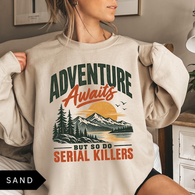 May include: A sand-colored crewneck sweatshirt featuring a graphic print with the text "ADVENTURE Awaits BUT SO DO SERIAL KILLERS". The design includes a mountain landscape, pine trees, a lake, and a sun in shades of green, orange, and black.