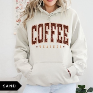 Puede incluir: Una sudadera con capucha beige con un gr&aacute;fico marr&oacute;n que dice "COFFEE WEATHER" con estrellas a cada lado del texto.