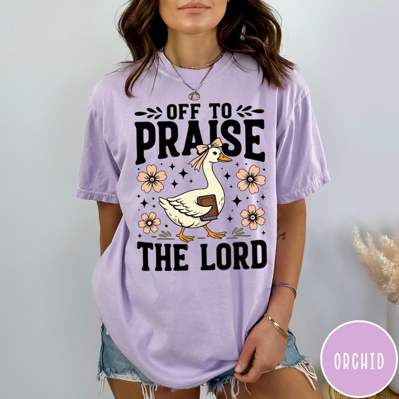 Puede incluir: Camiseta lila claro con el texto "OFF TO PRAISE THE LORD". El dise&ntilde;o presenta un ganso con un libro, rodeado de flores color melocot&oacute;n. La camiseta es de estilo casual y c&oacute;modo.