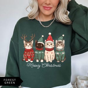 May include: Forest green sweatshirt featuring four cartoon cats dressed for Christmas. The cats wear sweaters and hats, with the text "Meowy Christmas" below. The cat on the left has antlers, and the others have hats.