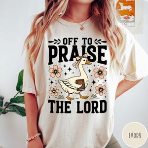 Puede incluir: Camiseta color marfil con un ganso sosteniendo un libro y el texto "OFF TO PRAISE THE LORD". El dise&ntilde;o incluye flores y estrellas color melocot&oacute;n. Es una prenda de vestir informal.