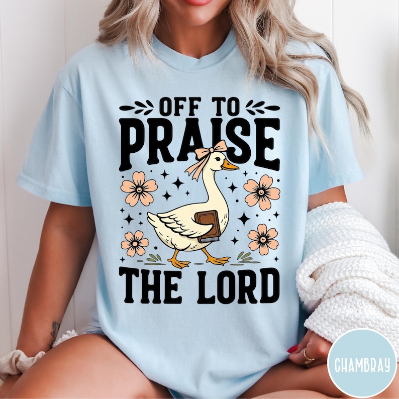 Puede incluir: Camiseta azul claro con el texto negro "OFF TO PRAISE THE LORD". En el centro, un ganso de dibujos animados con un lazo y un libro, rodeado de flores y estrellas color melocot&oacute;n. La camiseta es de estilo casual.