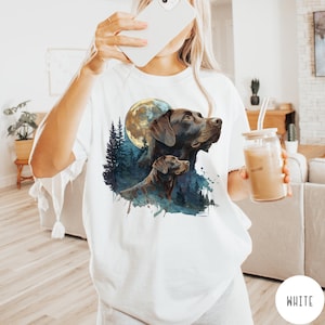 May include: White t-shirt featuring a watercolor illustration of two brown Labrador Retrievers with a full moon and forest background. The design has blue and green accents. The shirt is a casual style.