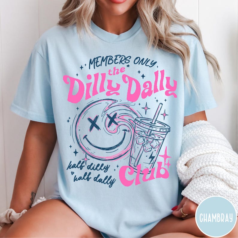 May include: Light blue t-shirt with a graphic design featuring the text "MEMBERS ONLY the Dilly Dally Club". The design includes a smiley face with an X for eyes, a drink, and the words "half dilly half dally".