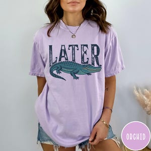 May include: A light purple t-shirt with the word "LATER" in dark blue block letters above a blue and green alligator illustration. The shirt is a relaxed fit and has short sleeves.