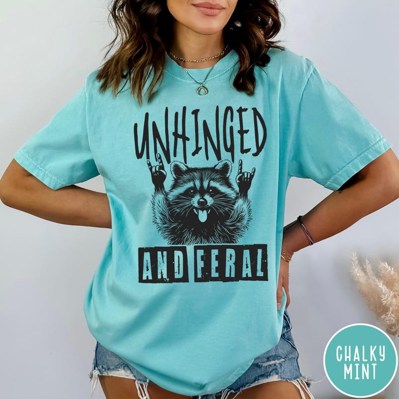 May include: A turquoise blue t-shirt with a black graphic of a raccoon with the text "Unhinged and Feral" printed on it.