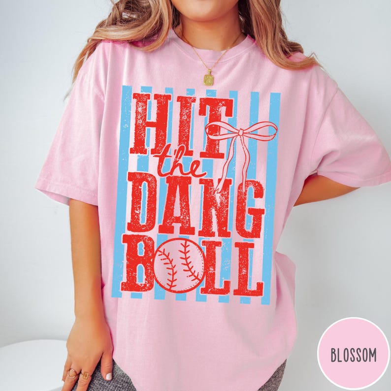 Pu&ograve; includere: T-shirt rosa chiaro con la scritta "HIT the DANG BALL" in lettere rosse. Il design include una palla da baseball e un fiocco, con strisce verticali azzurre. La maglietta &egrave; in stile casual.