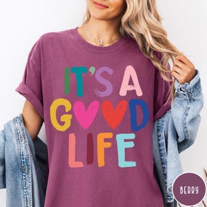 K&ouml;nnte beinhalten: Ein beerenfarbenes Kurzarm-T-Shirt mit dem Text IT'S A GOOD LIFE, der in einer mehrfarbigen Schriftart im Pinselstrich-Stil gedruckt ist. Die Buchstaben sind in den Farben Gr&uuml;n, T&uuml;rkis, Lila, Pink, Gelb, Rot, Blau und Orange gehalten.