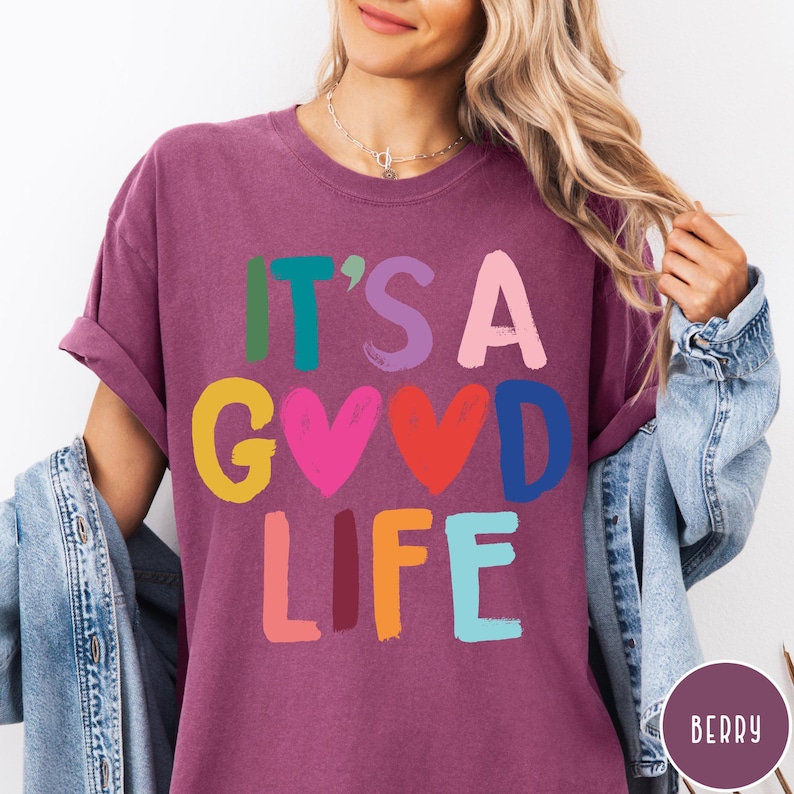 K&ouml;nnte beinhalten: Ein beerenfarbenes Kurzarm-T-Shirt mit dem Text IT'S A GOOD LIFE, der in einer mehrfarbigen Schriftart im Pinselstrich-Stil gedruckt ist. Die Buchstaben sind in den Farben Gr&uuml;n, T&uuml;rkis, Lila, Pink, Gelb, Rot, Blau und Orange gehalten.