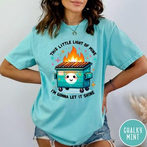 May include: A turquoise blue t-shirt with a graphic of a green dumpster on fire with a smiling face and the text "This little light of mine I'm gonna let it shine".
