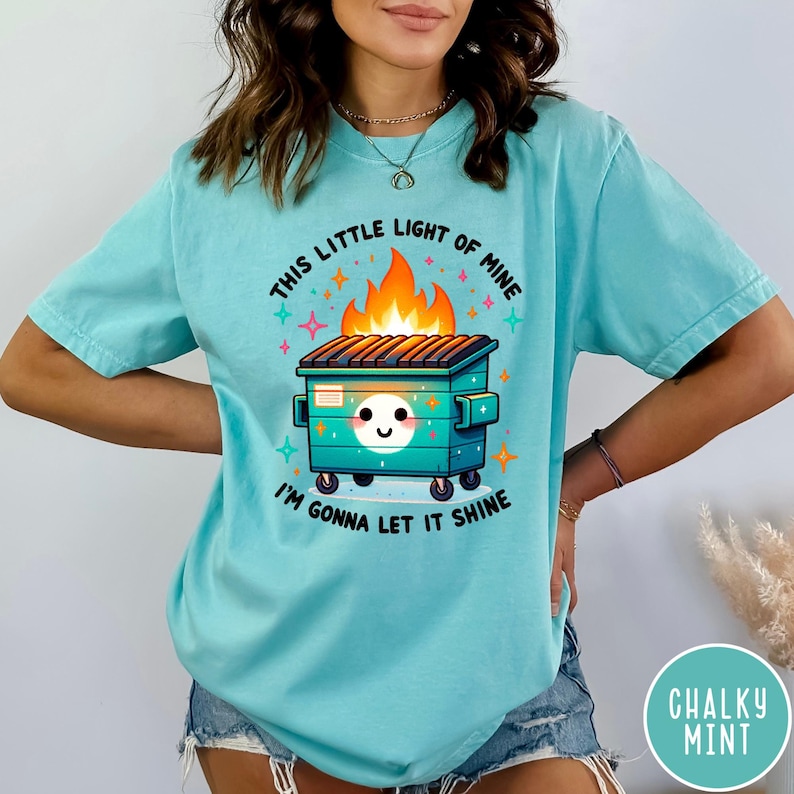 May include: A turquoise blue t-shirt with a graphic of a green dumpster on fire with a smiling face and the text "This little light of mine I'm gonna let it shine".