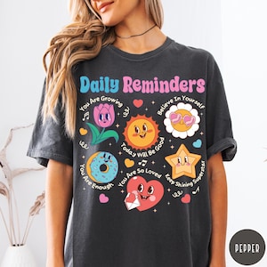 May include: Dark gray t-shirt with the text "Daily Reminders" in pink and blue. The shirt features colorful cartoon graphics of a flower, sun, star, donut, and heart with positive affirmations. The shirt is a comfortable, casual style.