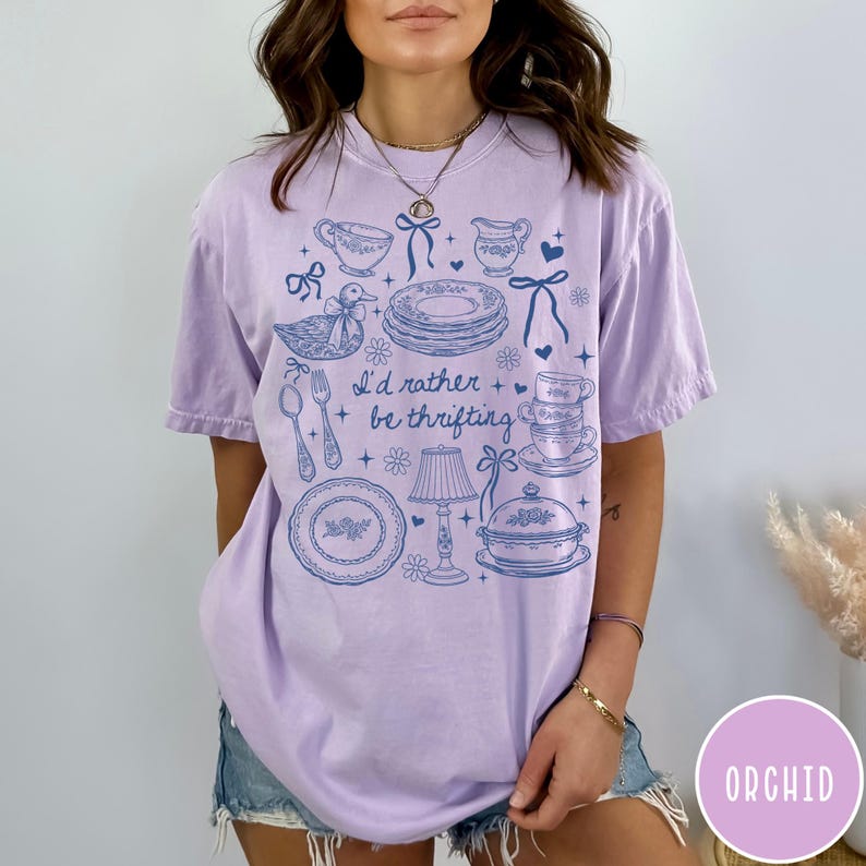 May include: A lilac t-shirt featuring a blue illustration of vintage kitchenware, such as plates, teacups, and a duck. The text "I'd rather be thrifting" is printed on the front of the shirt.
