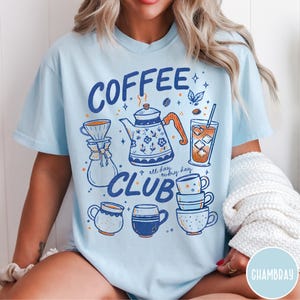 May include: Light blue t-shirt with a coffee-themed graphic. The design includes the words "COFFEE CLUB" and illustrations of a coffee pot, coffee cups, and a glass of iced coffee. The phrase "all day, every day" is also included.