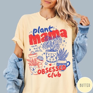 Puede incluir: Camiseta de color crema con un dise&ntilde;o gr&aacute;fico. El dise&ntilde;o incluye las palabras "plant mama" en rojo y azul, e ilustraciones de plantas en macetas. La frase "Obsessed Club" tambi&eacute;n est&aacute; en rojo. La camiseta tiene mangas cortas.
